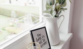 A cozy windowsill with a white kettle, a small plant, and a framed quote. In the background, a green garden is visible.