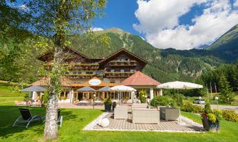 A charming hotel with a large garden and sunny terraces. In the background, green mountains and a blue sky can be seen.