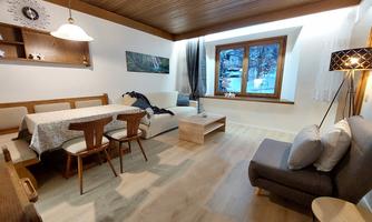 A cozy living space with a dining table, a couch, and large windows. The room is bright and features a rustic wooden ceiling style.