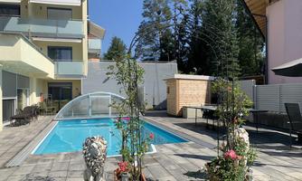 A beautiful swimming pool surrounded by plants and a sunny terrace. In the background, you can see a modern building.