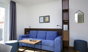 A modern living room with a blue sofa and a wooden table. Two pictures hang on the wall, and there is a bookshelf with a lamp.