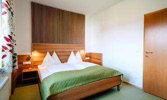 A modern hotel room with a double bed and wooden paneling. The walls are in light colors, and a window with colorful curtains provides daylight.
