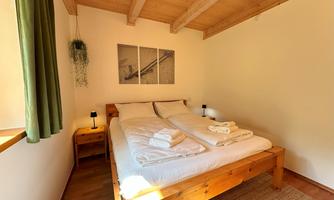A cozy bedroom with a wooden double bed and greenery at the windows. Pictures hang on the wall, and the room has a warm, inviting charm.