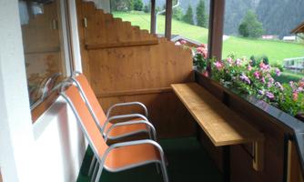 A cozy balcony with two metal chairs and a wooden tray. In the background, green meadows and mountains can be seen.