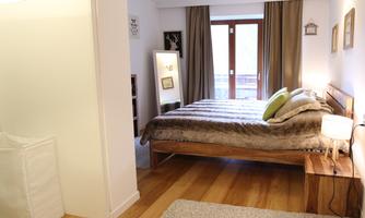 A cozy bedroom with a bed and wooden floor. Curtains hang in front of the windows, and a rug lies on the floor.