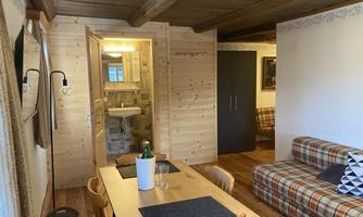 A cozy living space with a dining table, a couch, and an open kitchen area. The walls are made of wood, and the ambiance is rustic and inviting.