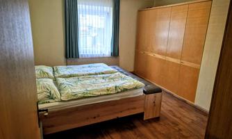 A simple bedroom with a double bed and a wooden dresser. The walls are painted light, and a window with curtains provides daylight.