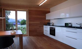 A modern kitchen with white cabinets and a wooden table. Large windows offer a view of the landscape.