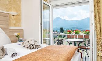 A comfortable hotel room with a view of the mountains. The balcony doors are open and colorful flowers are visible.