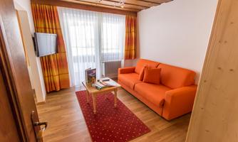 A cozy living room with an orange couch and a wooden table. Large windows let in plenty of light, and the floor is made of wood.