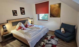 A cozy bedroom with a double bed and a blue armchair. The walls are subtly decorated, and a window lets in natural light.