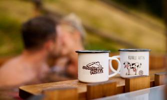 Two cups are standing on a wooden board while a couple kisses in the background. The cups have friendly designs and create a cozy atmosphere.