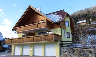 A modern house with a wooden balcony and a yellow paint. In the background, mountains and trees can be seen.