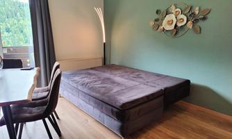 A modern room with a raised bed in dark fabric. A decorative flower hangs on the wall and the room has large windows overlooking greenery.