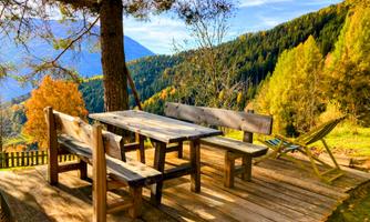 A cozy wooden table with benches is set on a terrace amidst colorful autumn trees. In the background, green mountains can be seen and the sky is blue.