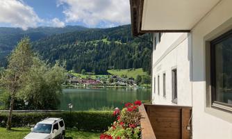 A peaceful landscape with a lake and green mountains in the background. In the foreground, there is a balcony with flowers and a car.