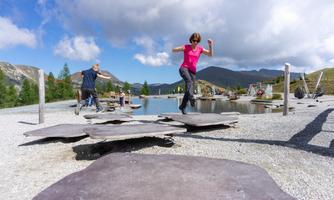 Two children are jumping over large stones in a play area by the water. In the background, mountains and a clear sky can be seen.