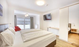 A modern bedroom with a large double bed and a television on the wall. Plenty of light comes in through the window, and there is a cozy armchair in the room.