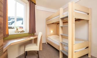 A bright room with a wooden bunk bed. There is a desk and a window facing outside.