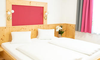A bright bedroom with two single beds and a wooden frame. In the room, there is a blooming plant and colorful curtains.