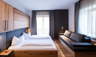 A modern hotel room with two single beds and a leather sofa. The walls are light, and wooden details create a cozy atmosphere.