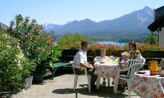 A family is sitting at a table on a terrace with flowers and a view of the mountains. The sun is shining and there is a clear blue sky.