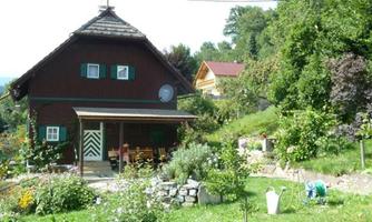 A rustic wooden house surrounded by a beautiful garden with many flowers and plants. In the background, additional houses and trees can be seen.