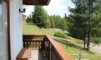 A balcony with a wooden railing and a small bench. In the background, green trees and a quiet street are visible.