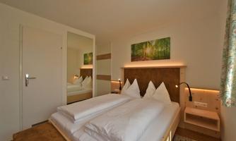 A cozy room with a double bed and white bed linens. A picture of a forest hangs on the wall, and there is a large mirror.