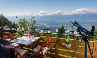 A viewing terrace with red chairs and a table displaying a bottle and glasses. In the background, an impressive mountain landscape stretches under a clear blue sky.