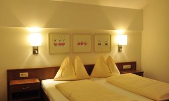 A cozy bedroom with two beds and decorative pillows. The walls are adorned with pictures, and warm light creates an inviting atmosphere.