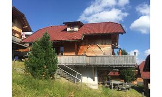 A cozy wooden house with a red roof stands on a meadow. In the background, there are more similar houses and a blue sky with some clouds.