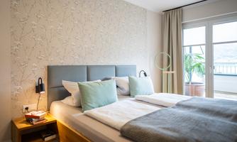 A modern bedroom with a comfortable double bed and bright pillows. Large windows offer a view of a balcony with plants.