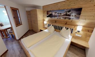 A cozy bedroom with a double bed and wooden paneling. On the wall hangs a picture of mountains, and there is a table and chairs in the background.