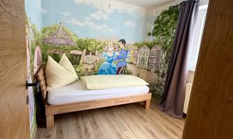 A cozy bedroom with a single bed and a colorful wall mural. The floor is made of wood and there is a modern lamp shade.