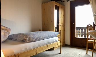 A simple room with a wooden bed and a wardrobe. A window with access to a balcony lets in a lot of light.