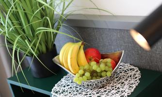 A fruit bowl with bananas, apples, and grapes is on a table. In the background, a green plant can be seen.