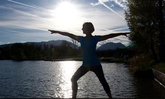 A person is doing a yoga pose by the shore of a lake. In the background, the sun is shining and the sky is clear.