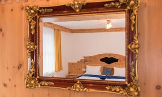 A mirror with an elaborate gold frame reflects a bright room. In the room, there is a bed and cozy wooden decor.