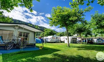 A campsite with green trees and caravans. In the background, there is a cozy accommodation and a clear sky.