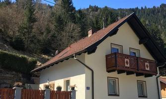 A cozy house with a red tiled roof and a small balcony. It stands in a mountainous area with trees in the background.