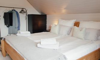 A cozy bedroom with a large double bed and bright ceilings. In the background, there is a clothes rack and a wardrobe.