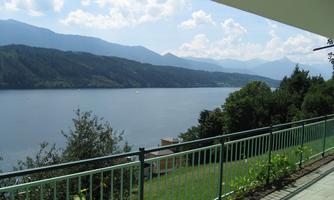 A peaceful lake with mountains in the background and a blue sky. A terrace with a railing offers a beautiful view of the landscape.
