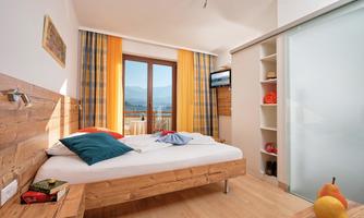 A modern bedroom with a large window that offers a view of the mountains. There is a bed with colorful pillows and a pleasant, bright atmosphere.
