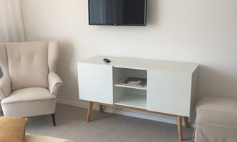 A modern living area with a cozy armchair and a white TV sideboard. A television hangs on the wall and there is a pillow on the floor.