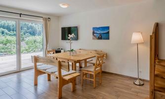 A bright dining room with a large wooden table and chairs. Through the window, you can see the green landscape outside.