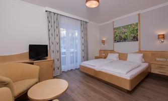 A modern hotel room with two single beds, a television, and a small table. Large windows provide plenty of daylight.