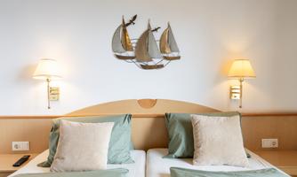 A cozy bedroom with two beds, decorated with pillows in soft colors. On the wall hangs a maritime decoration made of wooden ships, and there are two bedside lamps.