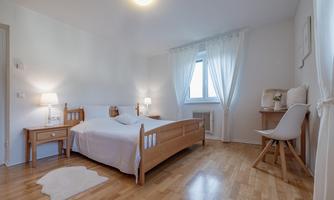 A bright bedroom with a wooden double bed and soft fabrics. There are nightstands, lamps, blinds, and a work area with a chair.