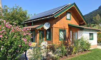 A charming wooden house with solar panels on the roof. The garden is green and blooming, surrounded by trees and shrubs.